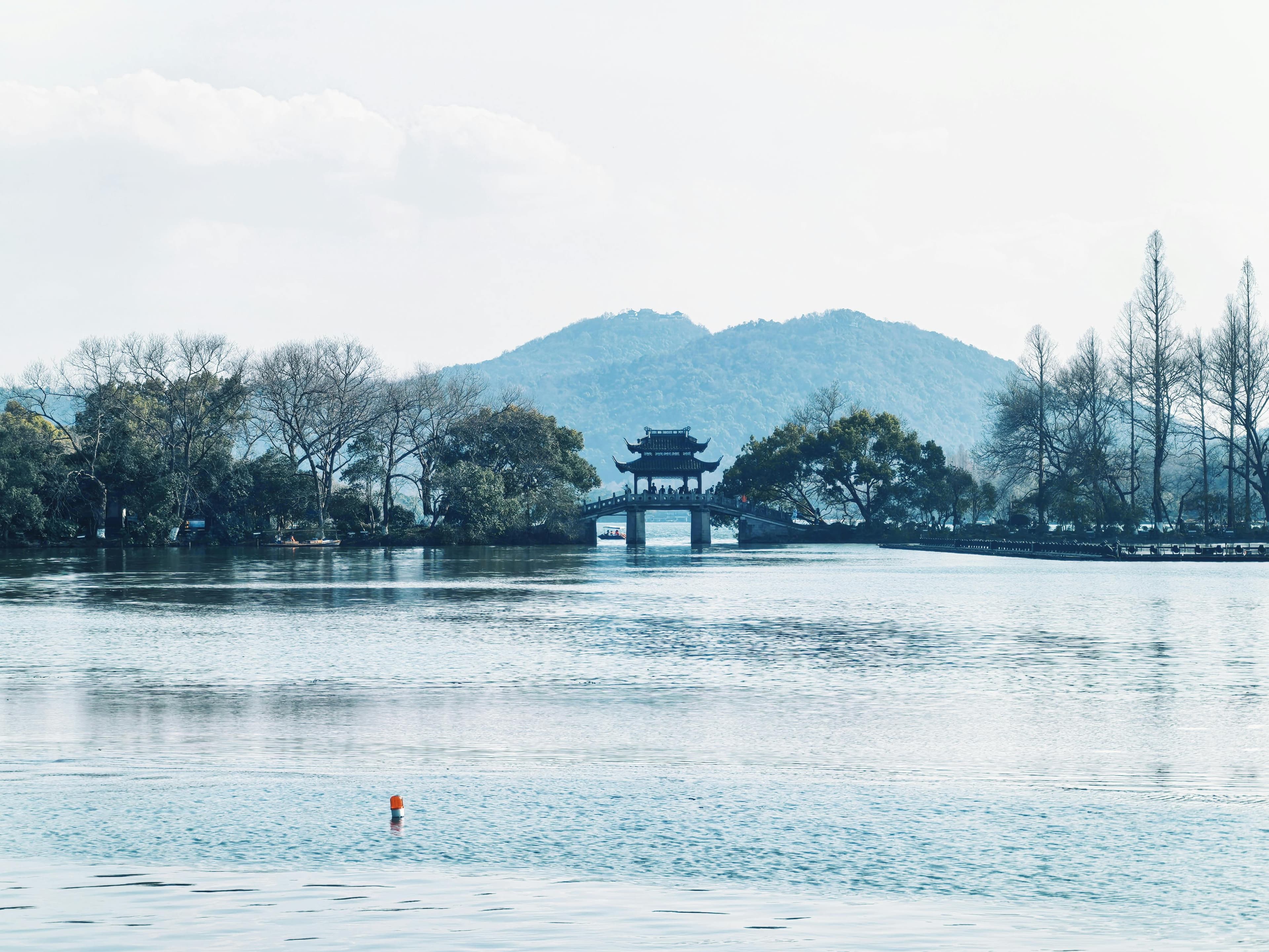West Lake in Hangzhou at sunrise with willow trees lining the shore, traditional pavilions, and misty hills in the background